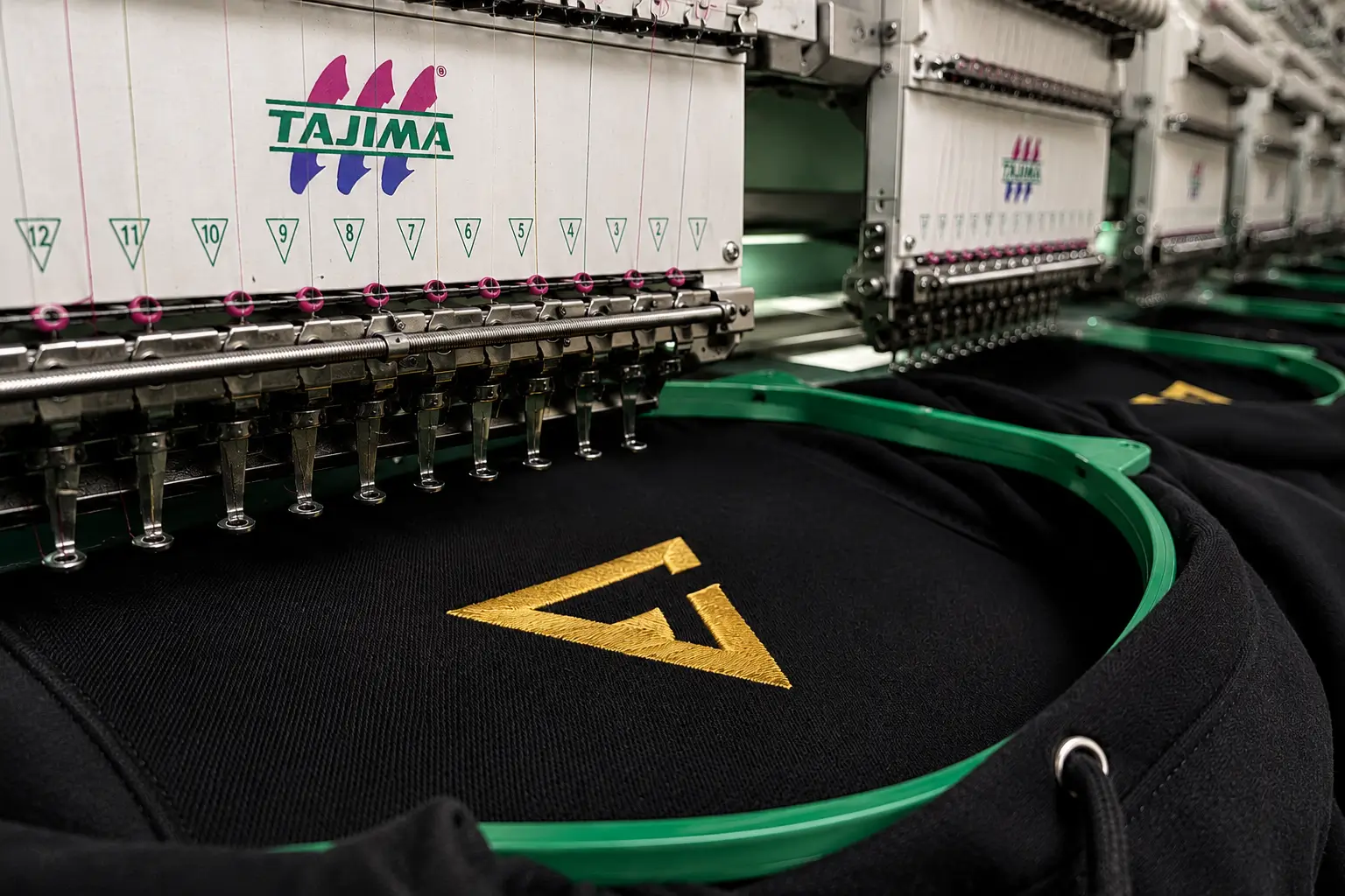 Custom Embroidery on a Tajima multi-head embroidery machine stitching a premium logo onto a black hoodie in a professional clothing manufacturing facility.