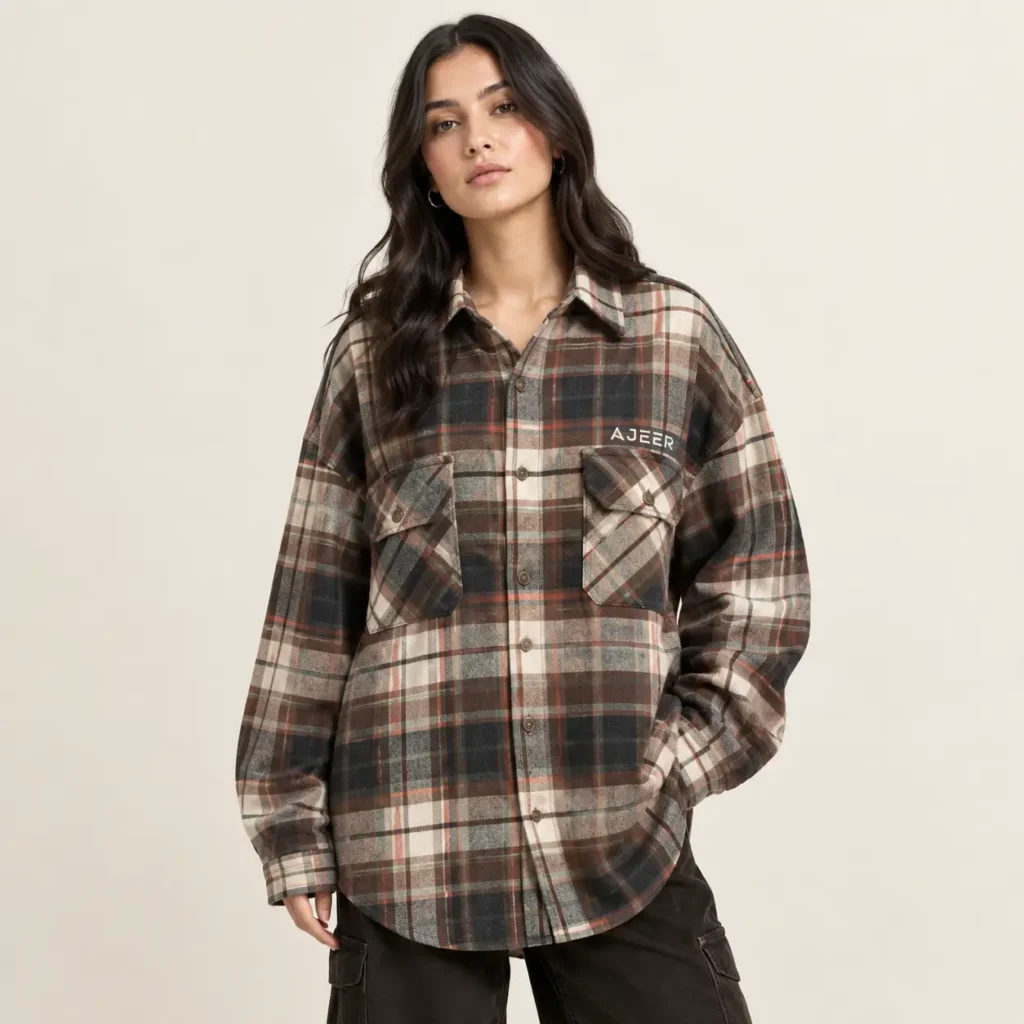 One adult female fashion model wearing a clearly oversized flannel shirt with relaxed proportions, dropped shoulders, wider body, longer sleeves, and a loose contemporary silhouette. Include a traditional collar, full button placket, button cuffs, and practical chest pockets. Plaid pattern with a sophisticated combination of brown, cream, charcoal, and muted accent colors. Small, AJEER INDUSTRIES logo on left chest.